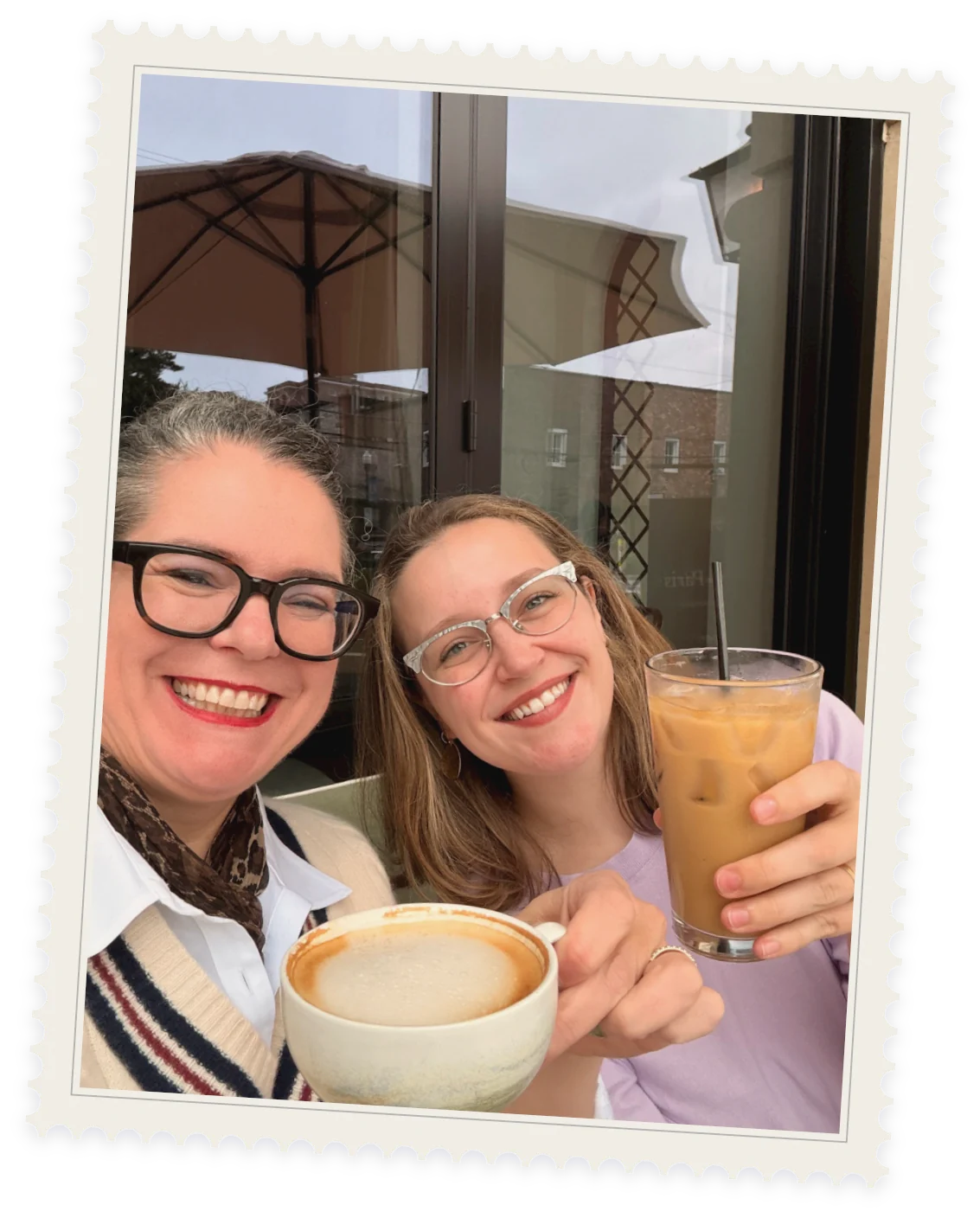 Laura and Kelsey at a café table outside, raising a cappuccino and an iced coffee to the camera, framed as a postage stamp.